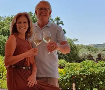 Un couple souriant profite d'une journée ensoleillée en plein air, verres de vin à la main. La verdure luxuriante et le ciel bleu clair créent une atmosphère relaxante et joyeuse pour leur célébration commune.