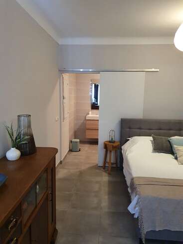 Cette chambre confortable comprend un lit bien fait, une armoire en bois, des vases décoratifs, du carrelage et une porte coulissante ouverte menant à une salle de bains moderne.