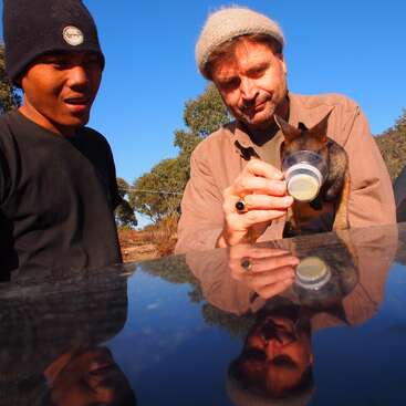 The image depicts two men standing beside a car, with one feeding a small animal from a bottle, set against a serene backdrop of trees and a clear blue sky.