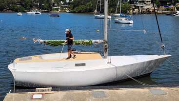 Un hombre está de pie en un velero blanco atracado junto al agua, sosteniendo una escoba. Otros barcos flotan cerca, con árboles y casas al fondo.