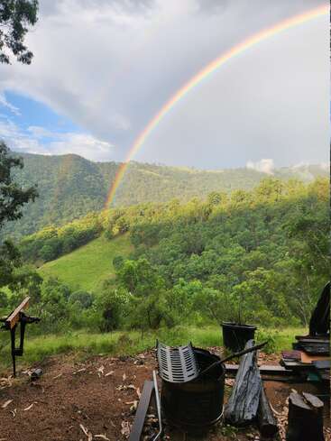 La imagen representa un paisaje sereno con un arco iris vibrante, exuberantes colinas verdes y árboles, con un montón de escombros en primer plano.