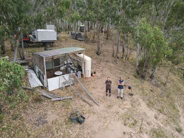 La imagen muestra a dos individuos de pie en una zona boscosa, rodeados por un pequeño cobertizo y varios objetos, con un camino de tierra que conduce al cobertizo.