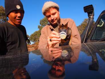 A imagem mostra dois homens em pé ao lado de um carro, com um deles alimentando um pequeno animal com uma mamadeira, em um cenário sereno de árvores e um céu azul claro.
