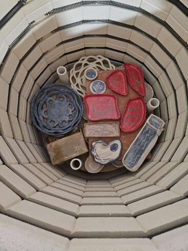Dentro de um forno de cerâmica, várias peças de cerâmica coloridas estão dispostas. As paredes internas do forno são revestidas de tijolos, prontas para a queima da cerâmica artesanal.