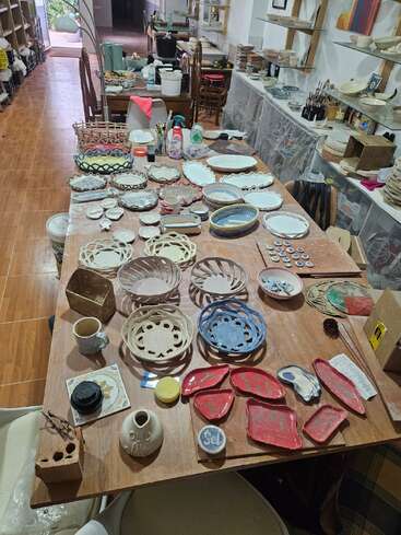 Uma grande mesa de madeira coberta com peças de cerâmica coloridas feitas à mão em um estúdio aconchegante, cercada por prateleiras, ferramentas, pincéis e materiais de cerâmica, prontos para serem esmaltados.