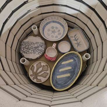 Dentro de um forno estão pratos de cerâmica pintados à mão, uma caneca e pequenos vasos cilíndricos. Cada peça exibe desenhos exclusivos, prontos para o processo final de queima.