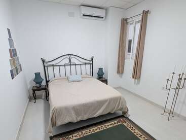 Il s'agit d'une chambre minimaliste comprenant un cadre de lit en métal, une literie beige, deux tables de chevet avec des lampes bleues, une fenêtre avec des rideaux et une décoration murale simple.