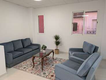 Cette image montre un salon simple et confortable avec des canapés bleus, un tapis coloré, deux plantes en pot, une table basse en verre et des murs blancs avec des fenêtres.