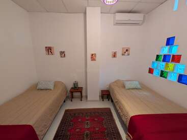L'image montre une chambre confortable avec deux lits simples, des couvre-lits beiges, de petites tables de chevet, des décorations murales, une fenêtre colorée, un climatiseur et un tapis à motifs.