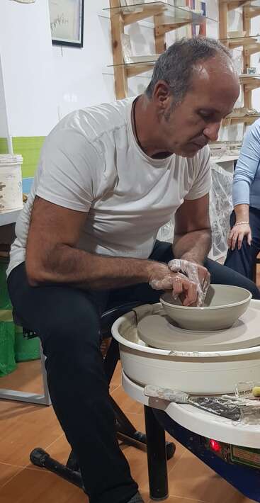 Un homme vêtu d'une chemise blanche est concentré sur le modelage de l'argile sur un tour de potier dans un atelier de céramique. Il est entouré d'étagères remplies de poteries et d'outils.