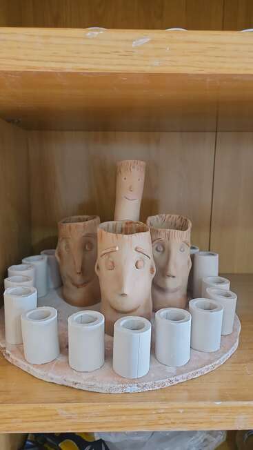 Four ceramic cups shaped like faces are arranged in a circle surrounded by smaller cylindrical clay pieces, all placed on a wooden shelf in a display.