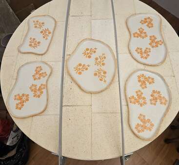 This image shows five irregularly shaped ceramic plates with orange floral patterns, placed on a round kiln shelf. Metal bars cross vertically over the plates.