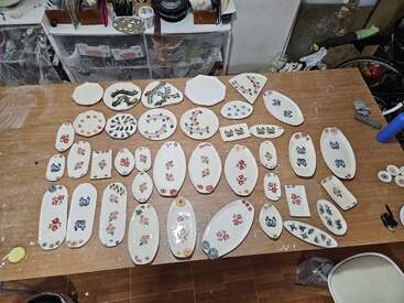 The image shows a wooden table covered with various handcrafted ceramic plates and trays, decorated with floral and animal patterns, organized neatly in several rows.