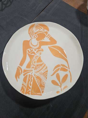 A white ceramic plate features an orange stencil-style art of a woman in traditional attire with jewelry and a plant motif, set against a dark background.
