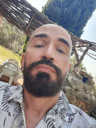 Un homme barbu au crâne rasé prend un selfie en gros plan en plein air. Il porte une chemise à fleurs et est assis près d'une structure rustique en bois, d'un mur en pierre, de chaises et d'arbres.