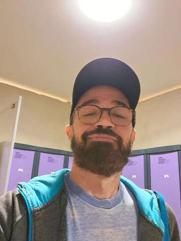 Un homme avec une barbe, des lunettes et une casquette sourit légèrement. Il porte un sweat à capuche et se tient devant des casiers violets à l'intérieur.