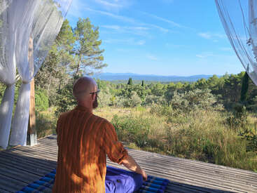 Un homme est assis les jambes croisées sur une terrasse en bois, entouré par la nature et des rideaux flottants, regardant paisiblement le paysage pittoresque, rempli d'arbres, sous un ciel bleu clair.