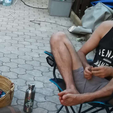 Eine Person sitzt barfuß in einem blauen Klappstuhl auf einer gepflasterten Fläche und trägt Shorts und ein ärmelloses Shirt. Ein Korb und eine Wasserflasche liegen in der Nähe.