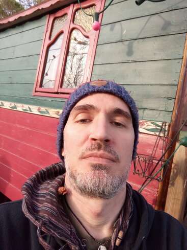 Ein Mann mit grauem Bart und blauer Strickmütze macht ein Selfie vor einem bunten Holzhaus mit rot und grün gestrichenen Wänden und Fenstern.