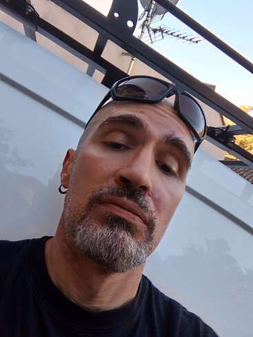 Ein Mann mit kurzen Haaren und einem grauen Bart posiert für ein Selfie im Freien. Er trägt eine Sonnenbrille auf dem Kopf, ein schwarzes Hemd und einen silbernen Ohrring.