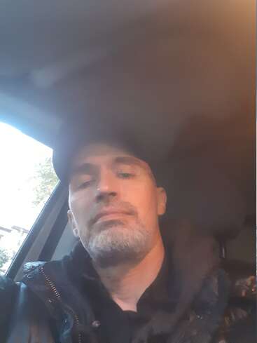 Ein Mann mit einem kurzen Bart und Schnurrbart, der eine dunkle Jacke und eine Mütze trägt, sitzt in einem Auto. Das Foto ist leicht schummrig und lässig.