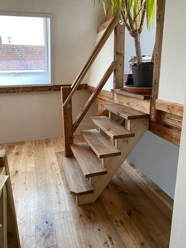 Dieses Bild zeigt eine kleine Holztreppe mit rustikalem Geländer, die zu einem Treppenabsatz mit einer Topfpflanze führt. Durch ein Fenster in der Nähe fällt Sonnenlicht ein.