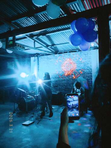 A band performs under blue stage lights and balloons. Someone records the scene on their phone, capturing the vibrant, artistic atmosphere with brick walls and projections.