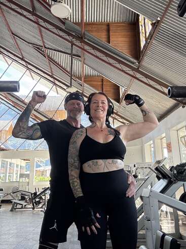 A smiling tattooed couple poses in a bright, industrial-style gym. Both flex their arms confidently, showcasing strength and camaraderie. Weightlifting equipment surrounds them. Empowered and happy.