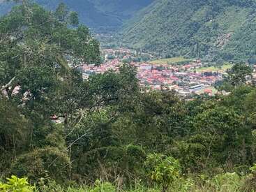 A lush, green forest frames a beautiful valley town with red-roofed buildings nestled among mountains, capturing a peaceful, scenic landscape from above.