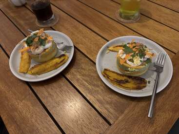 Two plates with savory food on a wooden table. Each has a stuffed bread roll topped with vegetables and herbs, served with grilled plantains. Two drinks nearby.