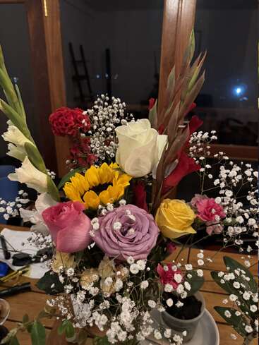 A vibrant floral arrangement featuring sunflowers, roses, carnations, gladiolus, baby's breath, and greenery sits on a wooden table indoors, illuminated by warm evening light.