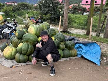 A man dressed in black squats and smiles in front of a large pile of green pumpkins outdoors, with motorcycles and trees in the background.