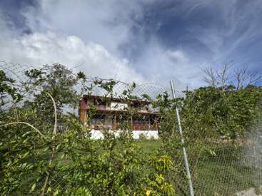 A two-story house with a balcony stands surrounded by lush greenery, viewed through a wire fence. Cloudy blue sky and overgrown plants dominate the peaceful scene.