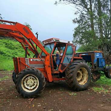 Das Bild zeigt einen Mann und zwei Kinder in einem roten Traktor mit einer blauen Maschine hinter ihnen, vor einem Hintergrund aus Bäumen und einem grasbewachsenen Hügel.