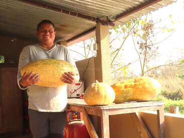 Un hombre sonriente está de pie en un porche con una calabaza enorme en las manos. Junto a él, sobre una mesa de madera, hay otras dos calabazas grandes bajo un cielo soleado.