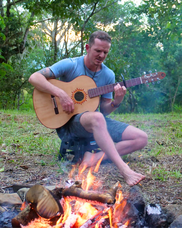 Ein Mann sitzt barfuß am Lagerfeuer und spielt in einer friedlichen Umgebung im Freien, umgeben von Bäumen und viel Grün, in der Abenddämmerung auf einer Akustikgitarre. Entspannte Atmosphäre.