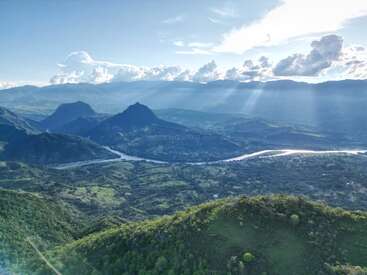 Eine atemberaubende Landschaft zeigt sanfte grüne Hügel, einen sich schlängelnden Fluss, hoch aufragende Berge und Sonnenstrahlen, die durch dramatische Wolken brechen und eine ruhige und malerische Szene schaffen.