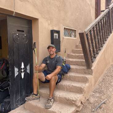 Un homme souriant en tenue de randonnée est assis sur des marches rustiques en pierre à l'extérieur d'un bâtiment, tenant des bâtons de randonnée, avec des sacs à dos et une porte ouverte visible à côté de lui.