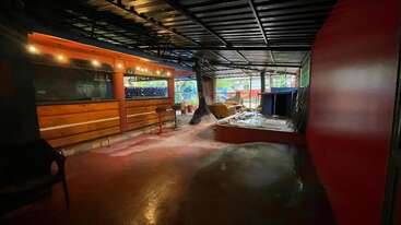 This image shows an indoor space under renovation, with red walls, benches, scattered construction materials, exposed ceiling, string lights, and some chairs, suggesting a future recreational or dining area.