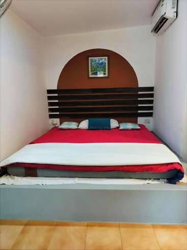 This cozy bedroom features a platform bed with red bedding, small pillows, wall-mounted artwork, air conditioner, brown semicircle accent wall, and light tiled flooring. Simple, modern design.
