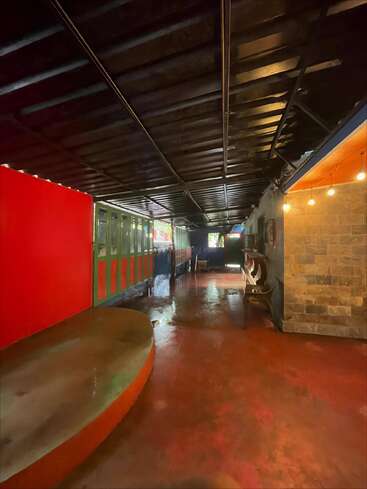 This image shows a dimly lit hallway with red and green walls, benches, exposed beam ceiling, stone wall, string lights, and natural light at the end.
