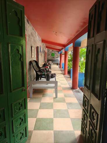 Open green doors reveal a tiled veranda with red columns, plastic chairs, and benches. Paintings hang on the wall. Lush greenery is visible outside.
