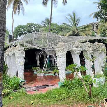 The image shows a unique outdoor pavilion structure supported by sculpted pillars shaped like faces, surrounded by tropical greenery, palm trees, and vibrant plant life.