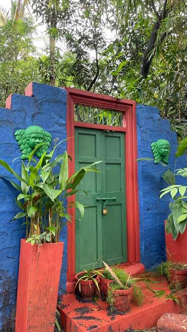 A vibrant green door set in a red and blue wall, decorated with green face sculptures, surrounded by lush plants and potted greenery outdoors.