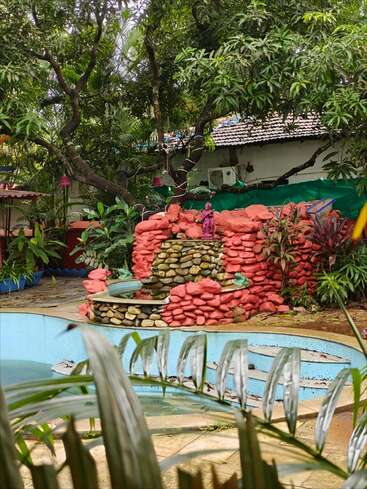 A serene garden scene with lush green trees, a dry blue swimming pool, stone decorations, red rocks, tropical plants, and a small purple statue centerpiece.