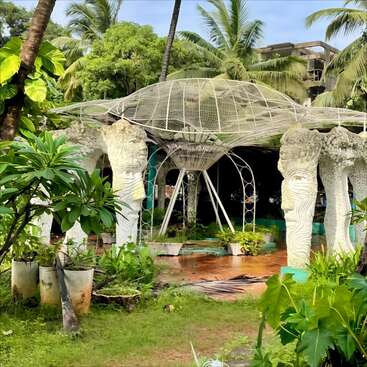 Cette image montre une structure de jardin unique avec des piliers sculptés ressemblant à des visages, entourés de plantes vertes luxuriantes et de palmiers, avec une canopée en fil de fer complexe au-dessus de la tête.