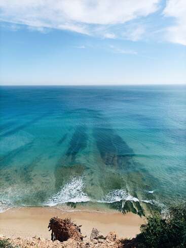 The image shows a stunning view from a cliff, overlooking a clear blue sea, gentle waves washing onto a sandy shore beneath a bright, partly cloudy sky.