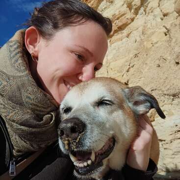 Eine Frau umarmt liebevoll ihren glücklichen, älteren Hund. Beide lächeln im Sonnenlicht vor einem Hintergrund aus Felsen und Sand und strahlen Wärme, Freude und Verbundenheit aus.