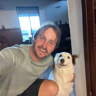 Um cara feliz tira uma selfie alegre com seu cachorrinho branco e fofinho dentro de casa, os dois sorrindo para a câmera. O quarto tem uma iluminação aconchegante e uma decoração aconchegante.
