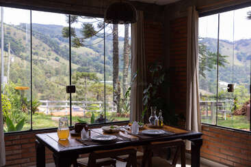A cozy dining area with a set table and juice overlooks lush, green hills and mountains through large glass windows, creating a peaceful, inviting atmosphere.
